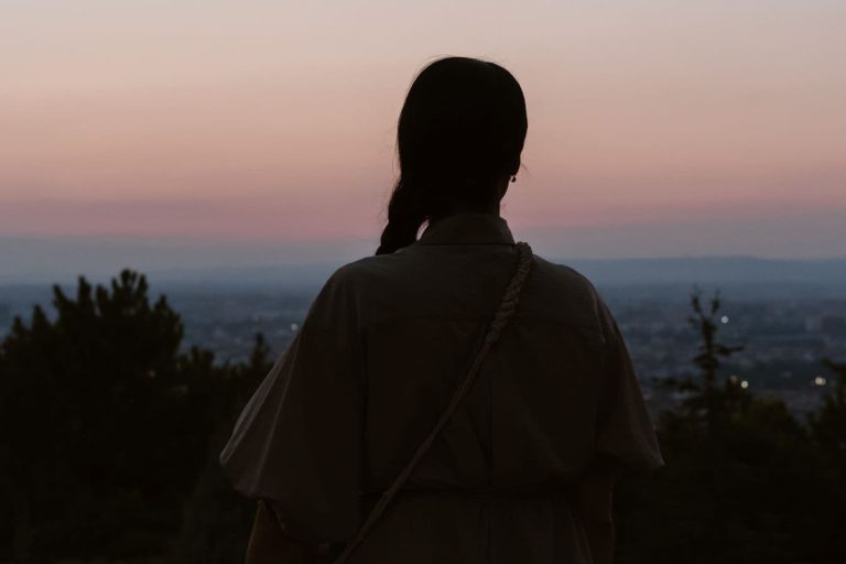 Naisen siluetti takaapäin kuvattuna. Naisen edessä alaviistossa kaupunki.